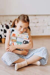 Adorable young girl hugging a stack of picture books while sitting on a wooden floor indoors.